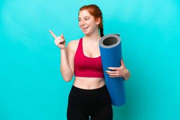 Young sport reddish woman going to yoga classes while holding a mat isolated on blue background...