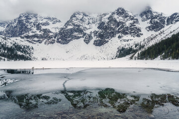 Frozen lake Morske Oko surrounded by stunning mountains in Tatra national park, Poland. Dramatic early spring landscape with snow, roks, clouds and reflection in clear water, outdoor travel background - 733811145