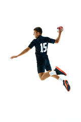 Full-length side view image of young man in dark uniform, handball player in motion, in a jump with ball during game, training over white studio background. Concept of sport, tournament, competition