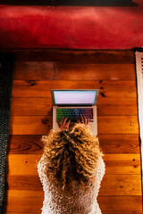 Adult woman with blonde curly hair work on a laptop computer on the wooden floor in the room - hotel or home worker technology and connection concept