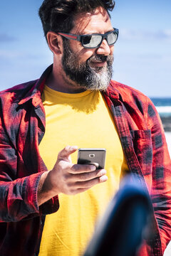 Travel and technology concept with portrait at handsome adult man with beard use modern phone outside his car with blue sky and sandy beach in background - roaming internet connection concept