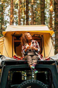 Travel and wanderlust lifestyle concept with happy lonely adult woman sit down on the roof tent car vehicle with wood forest in background enjoying nature and outdoors vacation