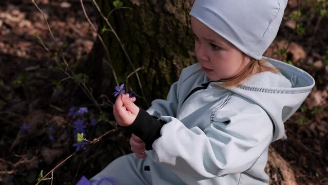 Little girl in violet boots and blue overall sitting under the tree in spring forest