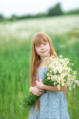 Portrait of a little smiling girl in a blue dress on a chamomile field with a bouquet of daisies on a sunny summer day