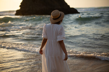 a young girl in a white dress walks on the beautiful ocean shore with sunset light and beautiful...