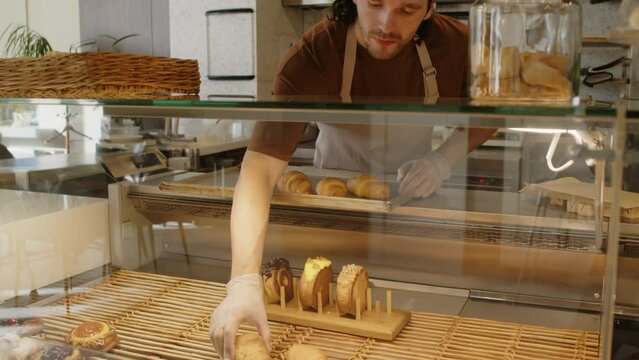 Tilt down shot of man in gloves and apron putting fresh croissants in glass display counter for sale in bakery