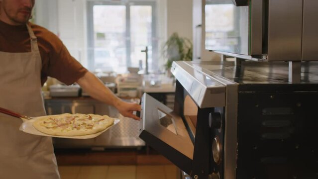Chef taking baked pizza from hot oven with peel and placing it on table in open kitchen while working in pizzeria