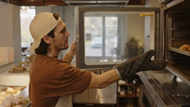 Young male baker in apron and mitt opening oven and taking tray with hot croissants while cooking in open kitchen in cafe