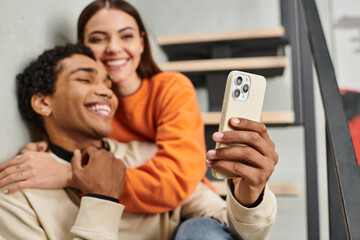 cheerful diverse couple sitting on stairs and smiling while taking selfie on smartphone in hostel