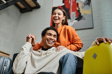 excited woman holding hands with her african american boyfriend near luggage in hostel room