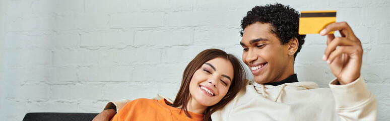 Cheerful diverse couple looking at a credit card while cozily reclining on living room sofa, banner - 733867950
