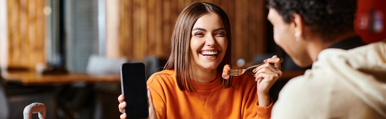 happy woman in orange sweater happily showing her phone to black boyfriend during meal, banner