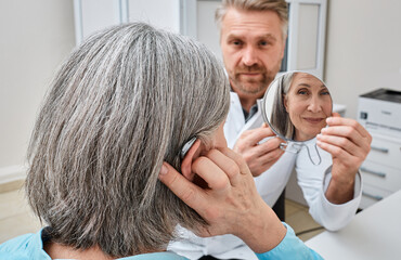 Senior woman trying on a hearing aid while looking into a mirror held by a hearing aid specialist...