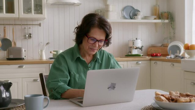 elderly woman sits at table in kitchen in morning with laptop. put on eyeglasses, types on keyboard looks in screen, smiles. succeed in using computer and internet, feels pleased and comfortable