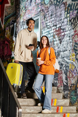 A cheerful diverse couple standing on stairs with colorful graffiti, one holding a yellow suitcase