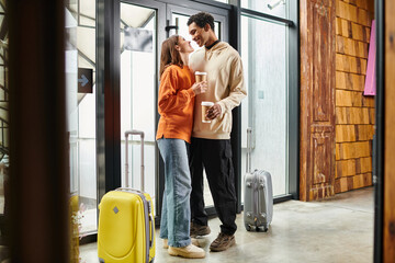 happy interracial couple with coffee embracing beside travel luggage in a modern entrance of hostel - obrazy, fototapety, plakaty