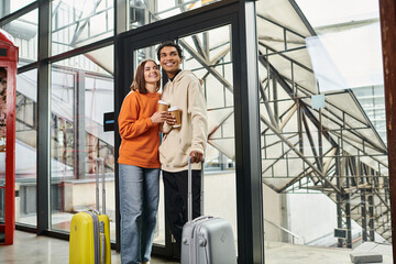 Fototapeta na wymiar Young diverse couple with travel luggage smiling and entering a modern hostel, holding coffee to go