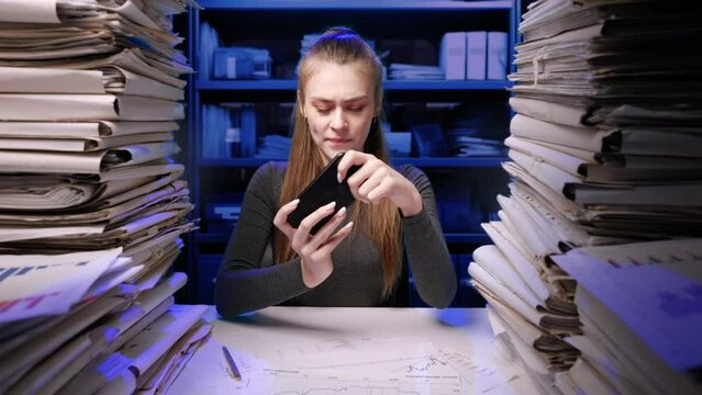 Exhausted young business woman office employee at the desk working late at night overloaded with paperwork. Female plays game on smartphone procrastinating work.