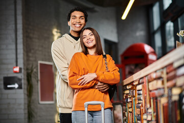 Satisfied diverse couple in casual attire embracing beside a counter of hostel near luggage