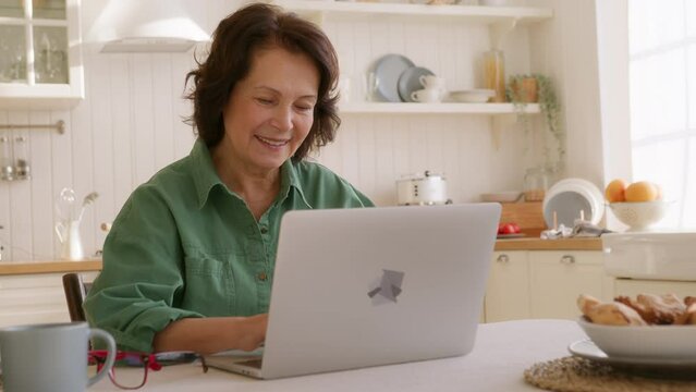 elderly woman sits at table in kitchen in morning with laptop. types on keyboard looks intently in screen, smiles. succeed in using computer and internet, feels pleased and comfortable