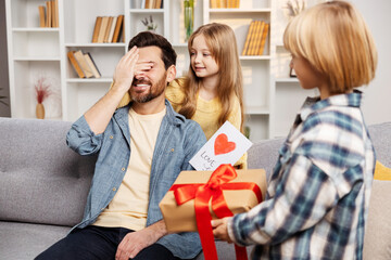 Kids surprise dad on couch with gifts, daughter playfully blinds him, engaging in 