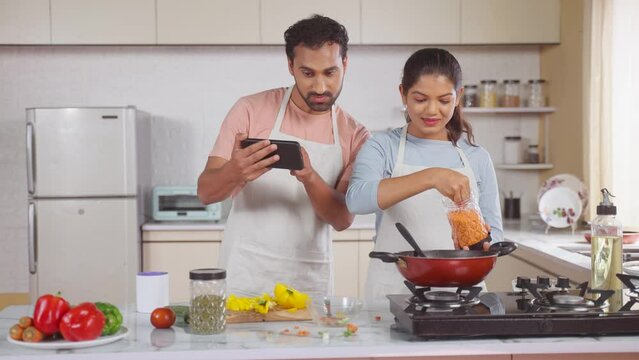 Joyful Indian couples cook or trying new food recipe by watching online videos from Mobile phone at Kitchen - concept of technology, relationship bonding and collaboration