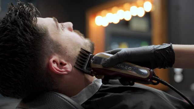 Barber shapes mans beard with electric trimmer in salon. Professional grooming, precise beard styling. Client relaxes, enjoys barbershop session. Master barber at work. Hair, beard care industry.