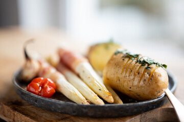 Fried white asparagus wrapped in bacon with herbed potatoes and tomatoes on black ceramic plate and wooden table. Short depth of field.