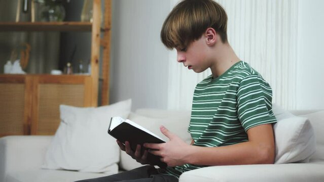 Relaxed educated young teenager man sitting on the sofa studying, reading a book, doing homework. Teenage guy boy reading holy book scripture. Religious christian kid child reading meditating praying