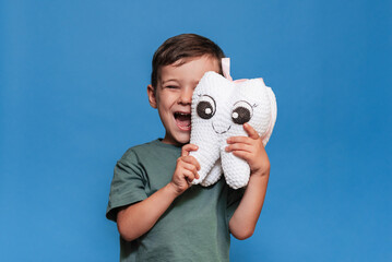 A smiling boy with healthy teeth holds a plush tooth on a blue background. Oral hygiene. Pediatric dentistry.