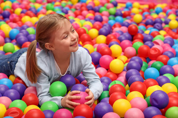 Fototapeta na wymiar Happy little girl lying on many colorful balls in ball pit, space for text