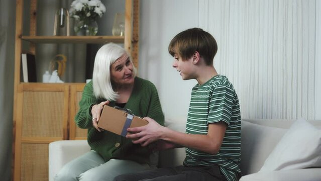 Happy excited grandson child teenager boy receiving present from loving old grandma embracing granny cuddling laughing happy to get gift box on birthday hugging grandmother make surprise to grandchild