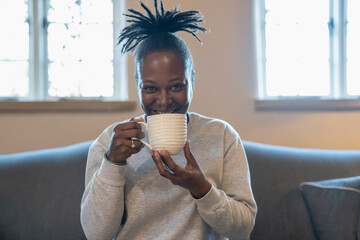 Happy woman with dreads drinking coffee while sitting on sofa - obrazy, fototapety, plakaty