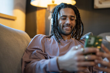 Man with dreads using phone and listening to music while relaxing on sofa