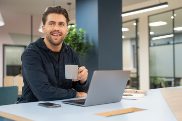 Smiling businessman working on laptop and drinking coffee in modern office