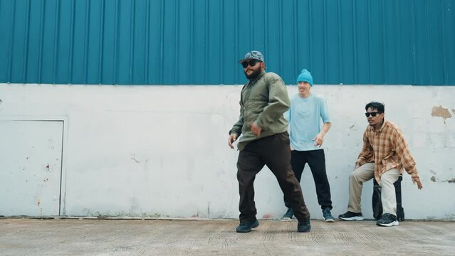 Skilled hipster doing cart wheel while multicultural friends encourage him. Hip-hop dancer moving foot step to music surrounded by group of hipster at wall near street. Outdoor sport 2024. hiphop.