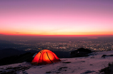 Orange tent on a snowy mountain slope against the background of the lights of the night city; romance and adventure concept