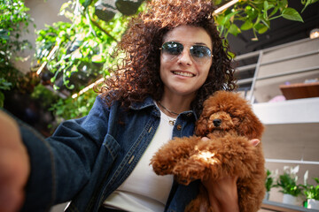 Selfie portrait brunette woman with curly hair holding small brown poodle dog and smiling indoors. Real happy female with pet