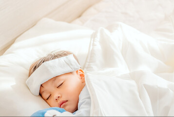 Little boy gets high fever lying on bed with cold compress, wet washcloth on forehead. Soft focus. Copy space.