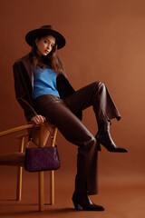 Fashionable confident woman wearing hat, blue jumper, faux leather suit blazer, pants, pointed toe ankle boots, with trendy purple  bag, posing on brown background. Full-length studio fashion portrait