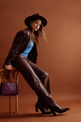 Fashionable confident woman wearing hat, blue jumper, faux leather suit blazer, pants, pointed toe ankle boots, with trendy purple  bag, posing on brown background. Full-length studio fashion portrait