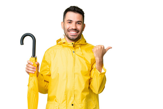 Young caucasian man with rainproof coat and umbrella over isolated background pointing to the side to present a product