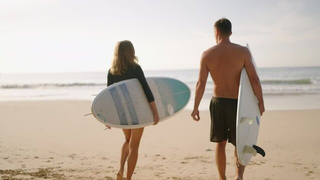 Athletic fit man, slender woman walk to ocean beach, carry surfboards. Young surfers couple dating. Boyfriend, girlfriend go surfing on sandy sea shore. Slim girl, guy with boards. Paradise coastline.