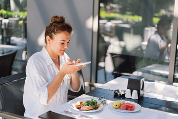 A woman uses a phone and eats lunch or breakfast outdoors in a cafe. Woman eating healthy food on a...