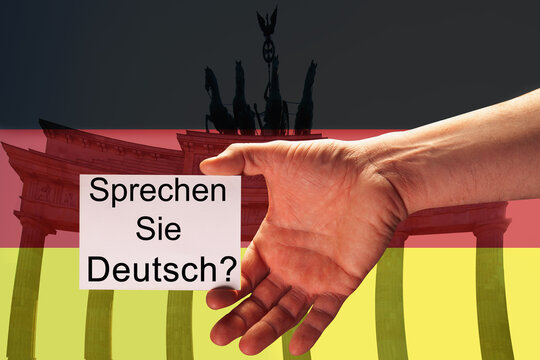 Closeup hand of young man holding white card with description: Sprechen sie deutsch? (Do You speak german?), written in german, against background of German flag and Brandenburg Gate