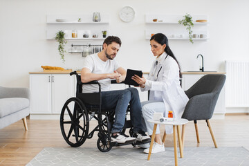 Full length shot of female brunette doctor checking up condition of bearded male patient at home. Nurse talking to mature man on wheelchair, showing examination results using modern tablet. - Powered by Adobe