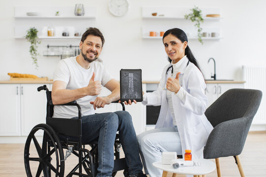 Portrait of young female nurse and mature man wheelchair user, showing x-ray scan of spine on modern tablet. Caucasian woman doctor checking up condition of mature bearded male patient at home.