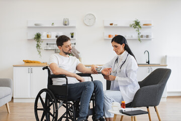 Attractive cardiologist telling patient with disability about all reasons of hypertension sitting at home. Adult man in wheelchair listening explanations about necessity of leading healthy lifestyle. - obrazy, fototapety, plakaty