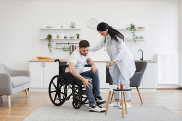 Focused female in doctor's coat supporting bearded male rising from wheelchair indoors. Health professional consulting man after back injury about physio therapy at home.