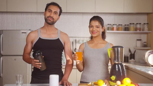 Portrait shot happy active couples with Protein shaker bottle and wife Orange juice looking camera at kitchen - concept of fitness routine, self care and togetherness
